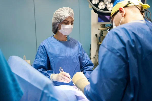 Surgeon operates the patient under a huge special lamp, the assistant has a surgical clamp in her hands