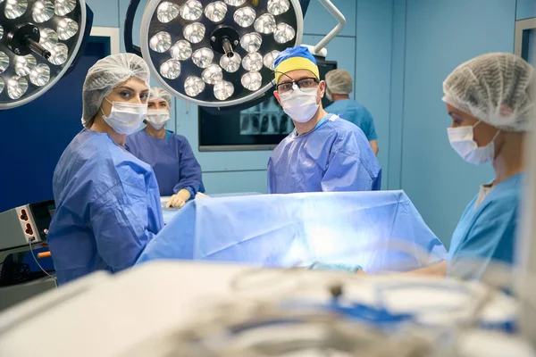 People in surgical gowns and protective masks stand in a modern operating room, powerful lamps above them