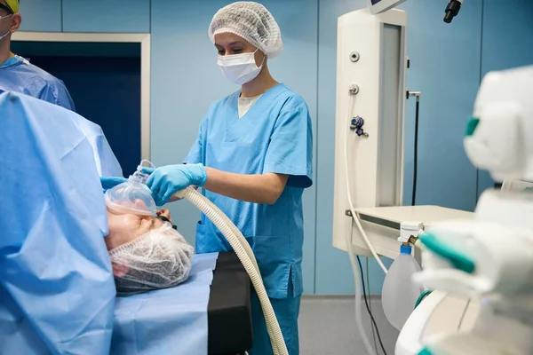 Female anesthesiologist holds a mask with anesthesia in front of the patient face, her colleagues work nearby