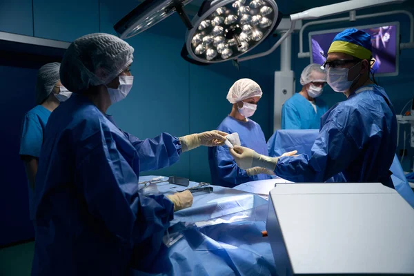 在手术过程中 女助手给了外科医生一个器械 一组医生在手术台上工作 — 图库照片