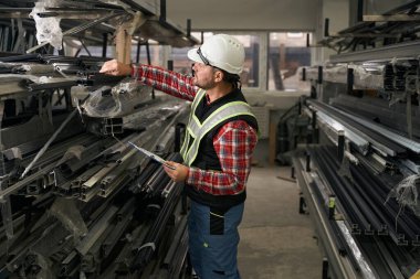 Koruyucu giysili, eldivenli ve kasklı bir adam metal profilli rafın yanında duruyor ve notlar alıyor.