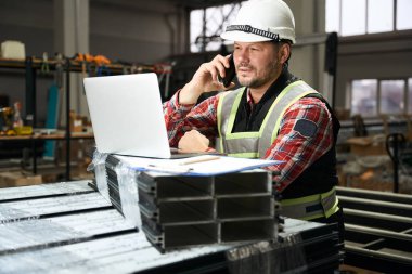 Koruyucu giysili, eldivenli ve kasklı bir adam atölyede dizüstü bilgisayarda çalışıyor, telefonla konuşuyor.