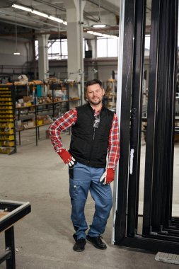 Profesyonel işçi koruyucu giysiler içinde, eldivenler atölyede duruyor, gülümsüyor ve kameraya bakıyor.