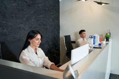 Genç bayan resepsiyonda oturuyor ve yanındaki monitöre bakıyor.