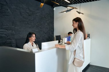 Gülümseyen yönetici masada oturup müşteriye bakıyor, kuaförde konuşuyor.