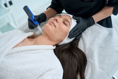 Koruyucu eldivenler ve palto tutma cihazı takmış dermatolog ve kuafördeki kadına akne tedavisi uyguluyor.