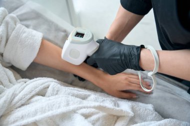 Koruyucu eldivenli kozmetik uzmanı yetişkin bir bayanın yanında duruyor ve eline lazerle saç aldırıyor.