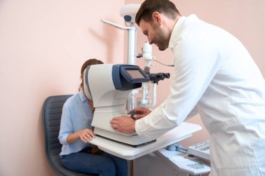 Beyaz önlüklü, gülümseyen sevimli doktor optometristin arka plan portresi tıp hastanesinde görme yetisini inceliyor.