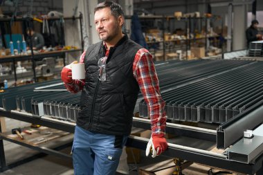Koruyucu giysili ve eldivenli bir adam çay bardağıyla fabrikada metal yapının yanında duruyor.
