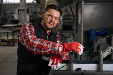 Koruyucu gözlüklü ve eldivenli bir erkek fabrikadaki çelik parçaları kesmek için ekipmanın yanında duruyor.