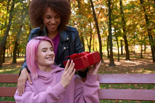 快乐的女士坐在木制公园的长椅上 从她微笑的女伴手中接过包装好的礼物 — 图库照片