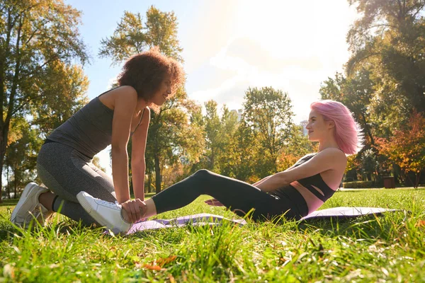 在她快乐的健身教练的帮助下 微笑着健康的女士在瑜伽垫上做仰卧起坐 — 图库照片