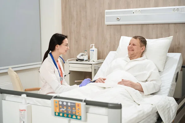 在现代病房里 躺在病床上的男性患者的侧面形象正在与女医生交谈 — 图库照片