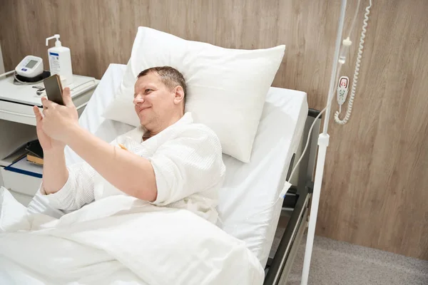 Portret Przystojnego Mężczyzny Leży Łóżku Medycznym Trzymając Telefon Komórkowy Nowoczesnym — Zdjęcie stockowe