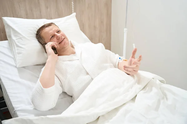 在医院里 一个面带微笑的勇敢的人躺在病床上 一边用手机交谈 一边近距离观察自己的近景 — 图库照片