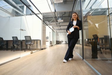 Ofis kıyafetleri içinde elinde dizüstü bilgisayarla kameraya bakan gülümseyen genç bayan.