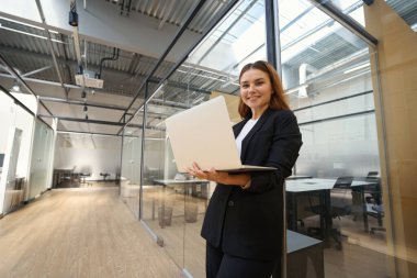 Ofis kıyafetleri içinde elinde dizüstü bilgisayarla kameraya bakarak gülümseyen genç bir kadın.