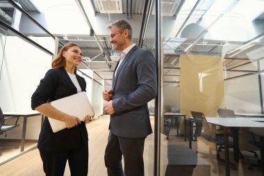 Ofis kıyafetleri içinde elinde dizüstü bilgisayarla duran ve bir adama bakan genç bir kadın.