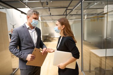 Ofis elbiseleri ve koruyucu maskeli genç bir bayan birlikte çalışıyor, dizüstü bilgisayarını tutuyor ve bir erkekle konuşuyor.