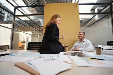 Kadın ve erkek, masanın yanında oturmuş, mimari planlarıyla ofiste sohbet ediyorlar.