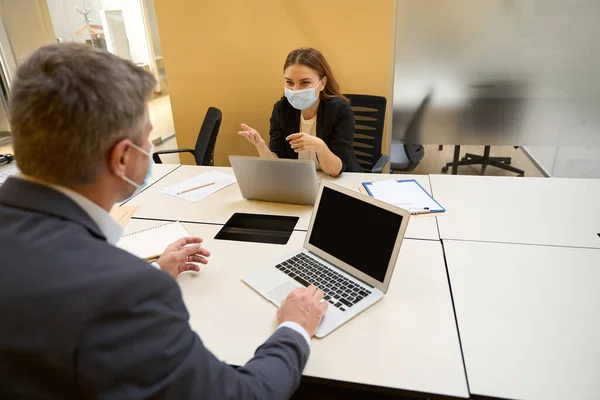 Workers in office clothes and protective masks sitting at the table with laptop and talking