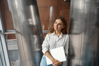 Ofiste dizüstü bilgisayarını tutan ve pencereye bakan mutlu genç bayan.