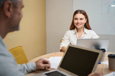 Dizüstü bilgisayarın yanındaki masada oturan kadın, ofis elbiseli adam karşıda oturuyor ve ona bakıyor.