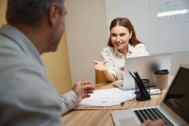 Dizüstü bilgisayarın yanındaki masada oturan kadın, ofis elbiseli adam karşıda oturuyor ve konuşuyor.