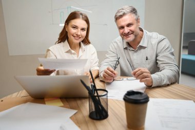 Dizüstü bilgisayarın yanındaki masada oturan genç bayan, ofis elbiseli adam, yanında oturuyor ve kameraya bakıyor.