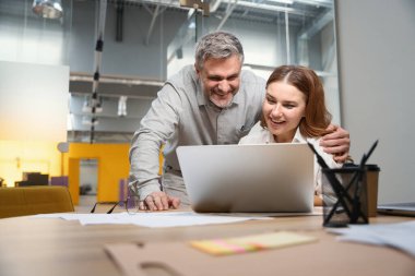 Dizüstü bilgisayarın yanındaki masada oturan bayan, ofis elbiseli mutlu adam, ekranın yanında duruyor ve ona bakıyor.