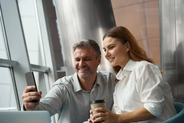 幸せな女性と男はノートパソコンの近くのオフィスに座って携帯電話を持って自撮り — ストック写真