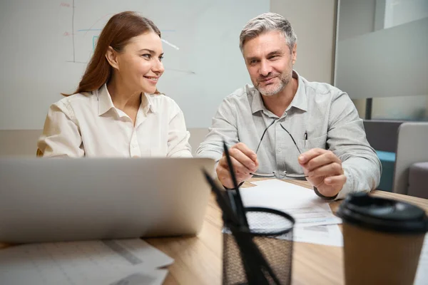 一个女人坐在笔记本电脑旁边的桌子旁 一个穿着办公室衣服的微笑男人坐在旁边 看着屏幕 — 图库照片