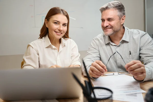 一个女人坐在笔记本电脑旁边的桌子旁边 一个穿着办公室衣服的微笑男人坐在旁边 看着她 — 图库照片