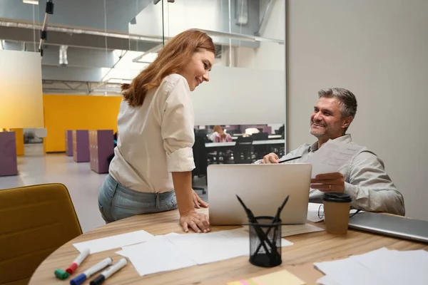 微笑的男人坐在笔记本电脑旁的桌子旁 穿着办公室衣服的女人站在旁边聊天 — 图库照片