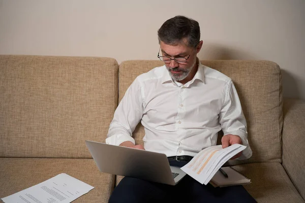 Male in business clothes sitting on the sofa, holding laptop and reading documents and working