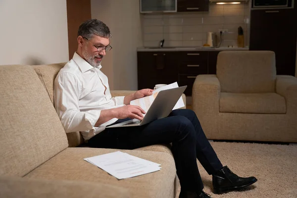 Man in business clothes sitting on the sofa, holding laptop and reading documents and working