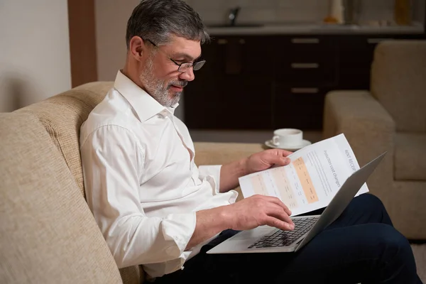 Man in business clothes sitting on the sofa, holding laptop and typing, reading documents, working