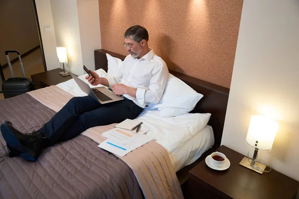 身穿商务服装的成年男性坐在床上 手持笔记本电脑和电话 — 图库照片