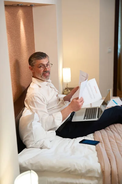 身穿商务服装的专业男性坐在床上 手持笔记本电脑和文件 — 图库照片