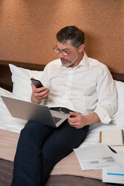 大人の男性ビジネス服をベッドの上に座って 携帯電話を保持し 画面上で見る — ストック写真