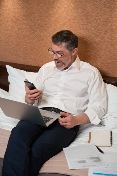 身穿商务服装的成年人坐在床上 拿着手机 看着屏幕 — 图库照片