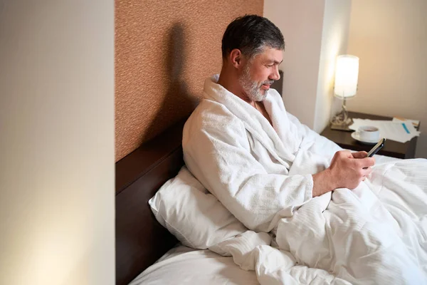 身穿浴衣的男性坐在床上 拿着手机 看新闻 — 图库照片