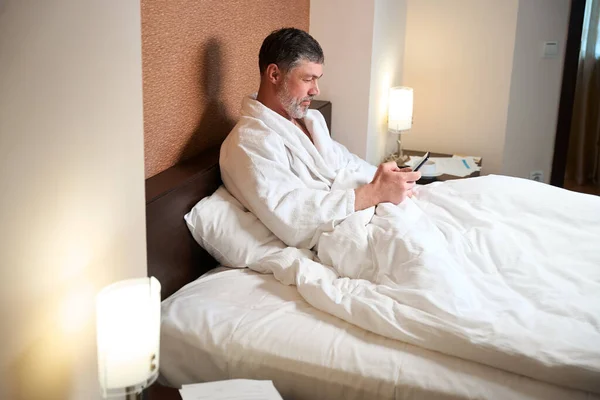 穿着浴衣的成年男性坐在床上 拿着手机 看新闻 — 图库照片