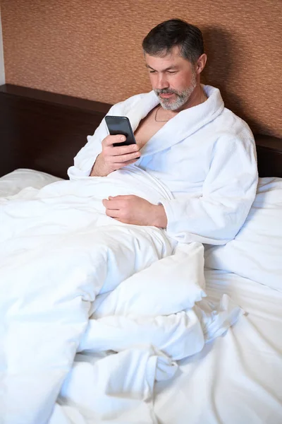 穿着浴衣的成年男性坐在床上 拿着手机 在酒店里看新闻 — 图库照片