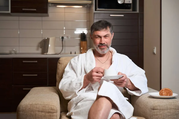 身穿浴衣的成年男性坐在厨房里 一边喝茶 一边喝酒 — 图库照片