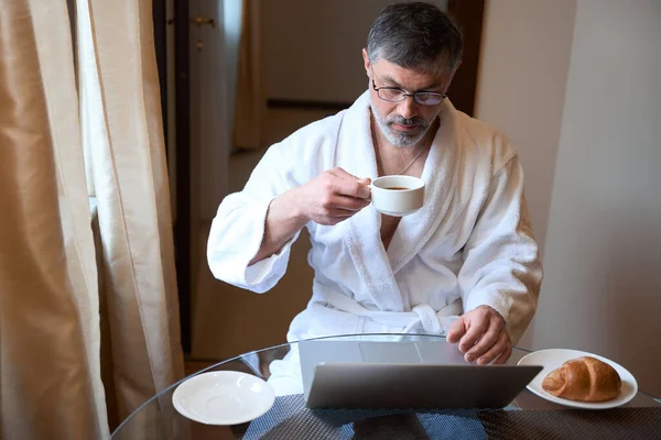 バスローブの大人の男はソファに座ってお茶を飲みながら仕事をしてる — ストック写真