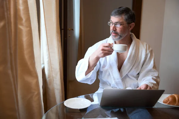 身穿浴衣的成年男性坐在沙发上 一边喝茶 一边喝酒和工作 — 图库照片