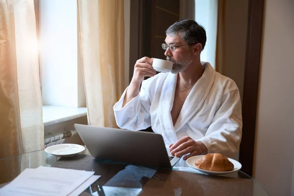 穿着浴衣的男人坐在沙发上 端着一杯茶 边喝酒边工作 — 图库照片