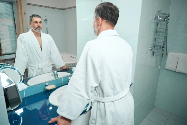Volwassen Man Witte Badjas Staan Badkamer Kijken Spiegel Onderzoekt Gezicht — Stockfoto