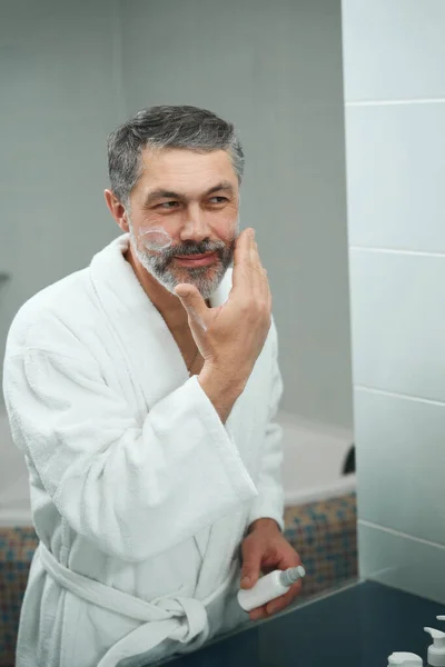 Homem Adulto Roupão Branco Banheiro Olhando Espelho Aplicando Creme Rosto — Fotografia de Stock